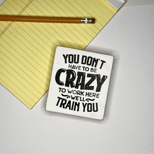 May include: A yellow legal pad with a pencil and a small white sign. The sign has black text that reads "YOU DON'T HAVE TO BE CRAZY TO WORK HERE WE'LL TRAIN YOU."