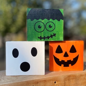 May include: Three wooden blocks with Halloween-themed faces. The top block is green with a Frankenstein face, the middle block is white with a ghost face, and the bottom block is orange with a jack-o'-lantern face.