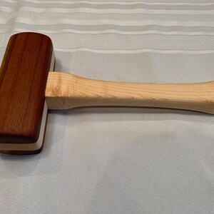 May include: A wooden mallet with a light brown handle and a dark brown head. The head is made of two different types of wood, creating a layered effect.