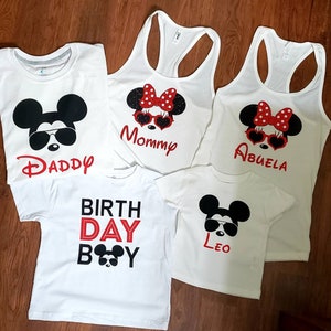 May include: Five white t-shirts with black and red Mickey Mouse designs. The shirts say "Daddy", "Mommy", "Abuela", "Birth Day Boy", and "Leo".