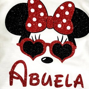 May include: A white t-shirt with a red glitter Minnie Mouse design. Minnie is wearing heart-shaped sunglasses and a red bow with white polka dots. The text "Abuela" is written in red glitter below Minnie.