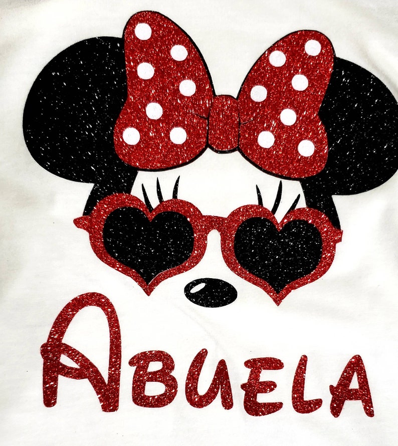 May include: A white t-shirt with a red glitter Minnie Mouse design. Minnie is wearing heart-shaped sunglasses and a red bow with white polka dots. The text "Abuela" is written in red glitter below Minnie.
