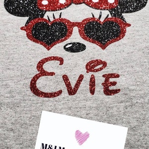May include: A gray t-shirt with a red glitter Minnie Mouse design with a red bow and sunglasses. The name "EVIE" is written in red glitter below the design. The t-shirt has a white tag with the text "M&I Magical Designs" and a website address.