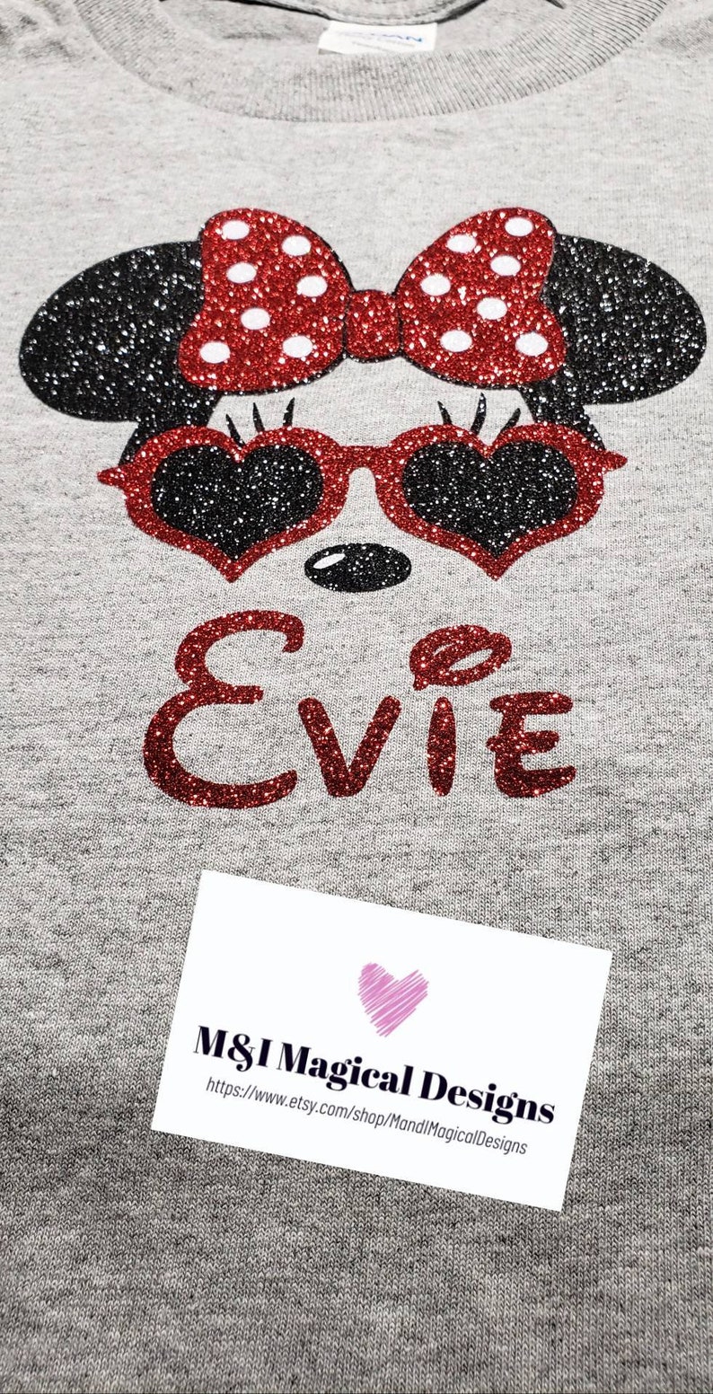 May include: A gray t-shirt with a red glitter Minnie Mouse design with a red bow and sunglasses. The name "EVIE" is written in red glitter below the design. The t-shirt has a white tag with the text "M&I Magical Designs" and a website address.