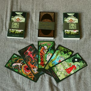 Könnte beinhalten: Eine Auswahl an Tarotkarten und -decks. Die Karten zeigen farbenfrohe Illustrationen mit einem dunkelgrünen Rand. Die Decks sind mit einem grünen Band umwickelt. Der Text "The simian" ist auf einem Deck zu sehen. Die Karten liegen auf einer grauen Oberfläche.