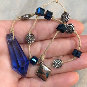 May include: A blue crystal pendant necklace with silver and blue beads. The necklace is made with a natural fiber cord.