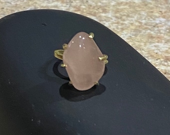 Puede incluir: Un anillo de oro con una piedra ovalada grande de color rosa claro. El anillo tiene un diseño simple con cuatro garras que sujetan la piedra.