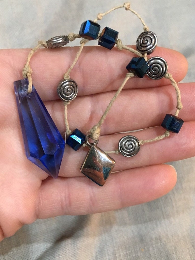 May include: A blue crystal pendant necklace with silver and blue beads. The necklace is made with a natural fiber cord.