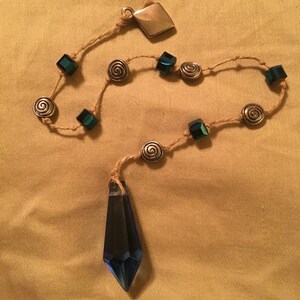 May include: A blue crystal pendulum with a brown cord and silver beads. The cord has green square beads and silver spiral beads.