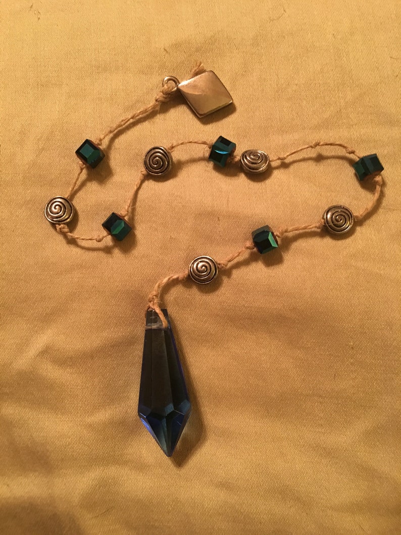 May include: A blue crystal pendulum with a brown cord and silver beads. The cord has green square beads and silver spiral beads.