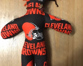 May include: A handmade doll with brown and orange yarn hair, wearing a black and orange fabric with the Cleveland Browns logo and text. The doll has button eyes and a brown cord necklace.
