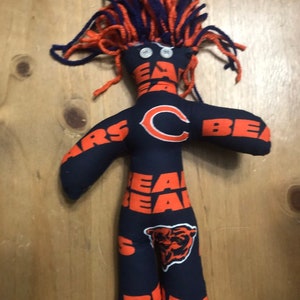 May include: A handmade rag doll with a blue and orange yarn hair. The doll is wearing a navy blue fabric with the Chicago Bears logo and the words "Bears" repeated on the fabric. The doll has two white buttons for eyes.