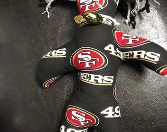 Może przedstawiać: Lalka z czarnej i czerwonej tkaniny z białymi wełnianymi włosami. Lalka nosi koszulkę San Francisco 49ers z logo drużyny i numerem 49. Lalka ma złoty wisiorek w kształcie piłki nożnej wokół szyi.