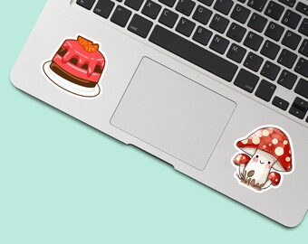 May include: Two vinyl stickers applied to a silver laptop. One sticker depicts a pink frosted cake with orange slices on top. The second sticker features a red and white spotted mushroom character with a smiling face and small arms.
