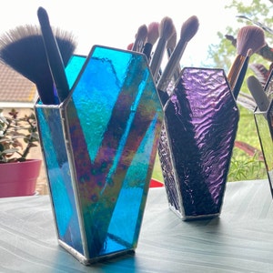 Puede incluir: Dos portacepillos de maquillaje de vidrio tintado en forma de ataúd. Uno es azul con acentos iridiscentes y el otro es morado con un patrón ondulado.