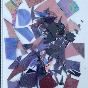 May include: A collection of various sized and shaped pieces of glass in shades of purple, blue, and black. The glass pieces are arranged on a white surface with a ruler in the background.