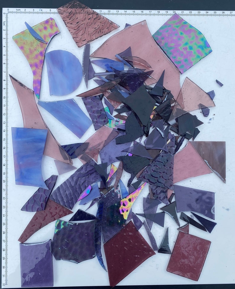 May include: A collection of various sized and shaped pieces of glass in shades of purple, blue, and black. The glass pieces are arranged on a white surface with a ruler in the background.