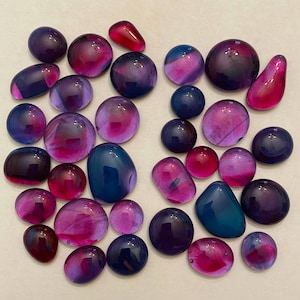 May include: A collection of polished glass stones in assorted shapes and sizes. The stones display shades of purple, pink, and blue, with some having a marbled effect. Some are oval, round, or teardrop-shaped, and all have a glossy finish.