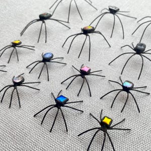 May include: A collection of small, handcrafted spider figurines. Each spider has a black wire body and legs, with colorful, iridescent glass or gem-like accents on their backs. The spiders are arranged on a textured, light gray surface.