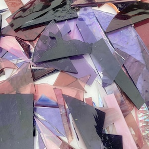 May include: A collection of purple and pink glass shards, some with iridescent qualities, arranged on a white surface. The glass pieces are of various sizes and shapes, creating a mosaic-like effect.
