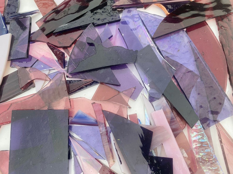 May include: A collection of purple and pink glass shards, some with iridescent qualities, arranged on a white surface. The glass pieces are of various sizes and shapes, creating a mosaic-like effect.