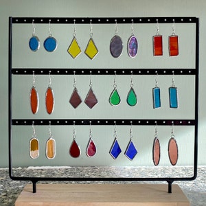 May include: A display stand with 15 pairs of stained glass earrings. The earrings are in various shapes and colors, including red, blue, green, yellow, and orange.