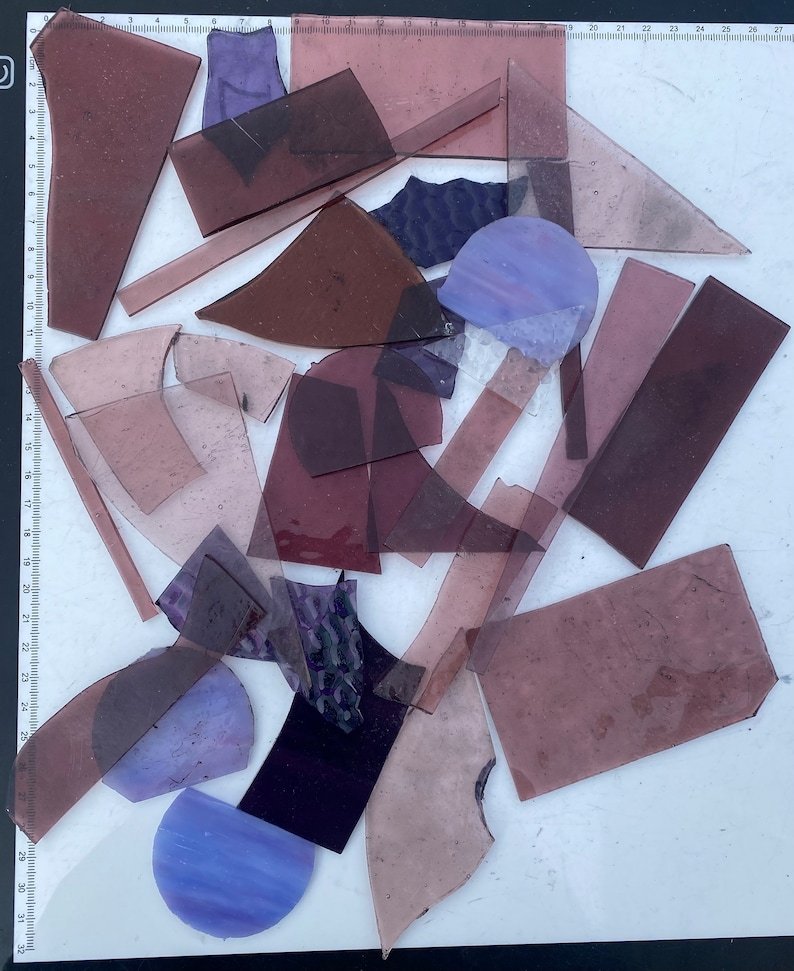 May include: A collection of purple and blue glass pieces in various shapes and sizes. The glass pieces are arranged on a white surface.