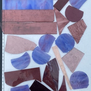 May include: Assortment of purple, brown, and beige glass pieces in various shapes and sizes. The pieces are arranged on a white surface with a ruler in the background.