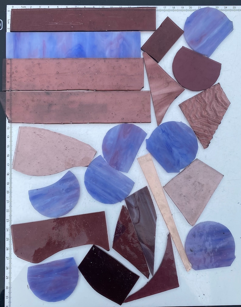 May include: Assortment of purple, brown, and beige glass pieces in various shapes and sizes. The pieces are arranged on a white surface with a ruler in the background.