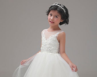 May include: A young girl wearing a white tulle dress with lace detailing and a crown. The dress has a sheer neckline and a full skirt. The girl is smiling and looking at the camera. The text "ERdress Shop" is visible at the bottom of the image.