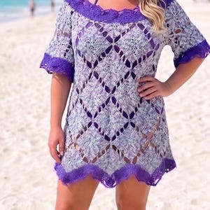 May include: A light purple and white crochet cover-up dress featuring a diamond pattern, short sleeves, and a solid purple scalloped trim along the neckline, sleeves, and hem. The garment is worn with a straw hat and brown sandals.