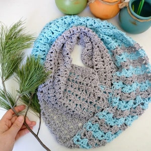 May include: A crocheted scarf in shades of grey and blue. The scarf is made with a delicate stitch pattern and has a fringed edge.