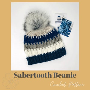 May include: A crochet beanie pattern featuring a blue, white and grey striped beanie with a faux fur pom pom. The pattern is called "Sabertooth Beanie" and is shown with a Caron X yarn colour card.