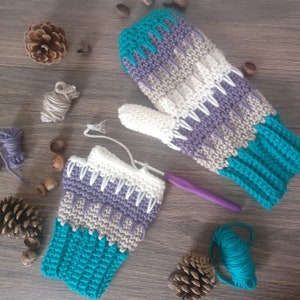 May include: A pair of teal, purple, white, and beige crocheted mittens. One mitten is complete and the other is in progress. The mittens are lying on a wooden surface with a crochet hook and yarn.