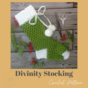 May include: Green and white crochet Christmas stocking with a white pom-pom on the top. The stocking is lying on a wooden surface with green leaves and red berries.