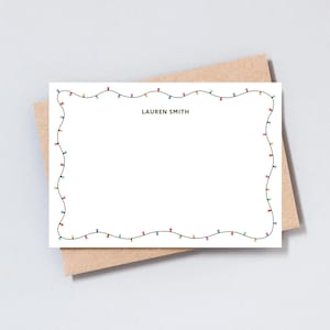 May include: A white stationery card with a border of colorful string lights. The name "LAUREN SMITH" is printed in black at the top. The card is placed on a brown envelope.