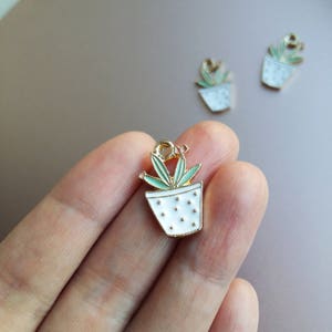 Pode incluir: Encanto de metal dourado com uma planta em vaso branco com folhas verdes. O vaso tem bolinhas brancas.