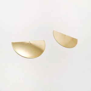 Könnte beinhalten: Zwei goldfarbene Halbkreis-Metallanhänger mit mattem Finish. Jeder Anhänger hat oben ein kleines Loch zum Auffädeln. Die Anhänger werden auf weißem Hintergrund dargestellt. Geeignet für die Schmuckherstellung oder Bastelprojekte.