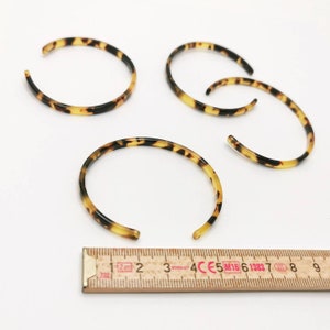 May include: Four open-ended bangles with a tortoiseshell pattern. The bracelets are a mix of brown, black, and yellow, and are arranged on a white surface. A ruler is placed near the bracelets.