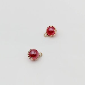 Könnte beinhalten: Zwei rote, runde Anhänger mit goldfarbenen Filigranfassungen und kleinen Schlaufen zum Befestigen an Schmuck. Die Anhänger haben eine glänzende Oberfläche vor weißem Hintergrund.