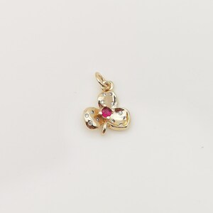 May include: A gold-coloured clover pendant with a small, round, red gemstone at its centre. The three clover leaves are adorned with small, clear gemstones. The pendant has a loop at the top for a chain.
