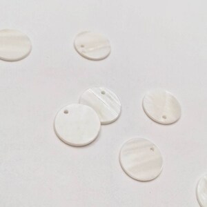 May include: A group of eight white, round, mother of pearl shell discs with a small hole in the center. The discs are scattered on a white surface.