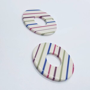 May include: Two white ceramic donut-shaped pendants with a hole in the center. The pendants are decorated with alternating stripes of green, pink, and blue.