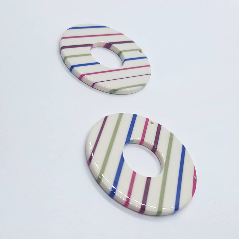May include: Two white ceramic donut-shaped pendants with a hole in the center. The pendants are decorated with alternating stripes of green, pink, and blue.