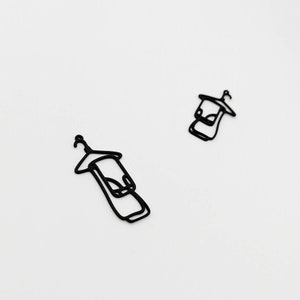 May include: Two black metal charm pendants shaped like a towel on a hanger. The pendants are cut out with a simple outline and are set against a white background. The pendants are small and delicate.