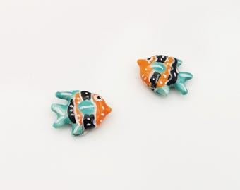 May include: Two ceramic fish figurines. Each fish has a turquoise body, orange head, and black and white striped details. The fish are small and appear to be handmade, with a glossy finish. They are set against a white background.