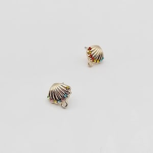 May include: Two silver-toned seashell stud earrings. Each earring features a fan-like design and is embellished with a row of colourful gemstones. The gemstones are in shades of red, yellow, green, and blue. The earrings have a post and loop for attachment.