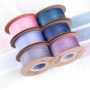 Puede incluir: Seis carretes de cinta de cuadros vichy en varios colores: rosa, verde azulado, azul marino, lavanda, morado y gris. La cinta está en carretes de cartón con la marca "ECCAIL". La cinta está dispuesta sobre una superficie blanca.