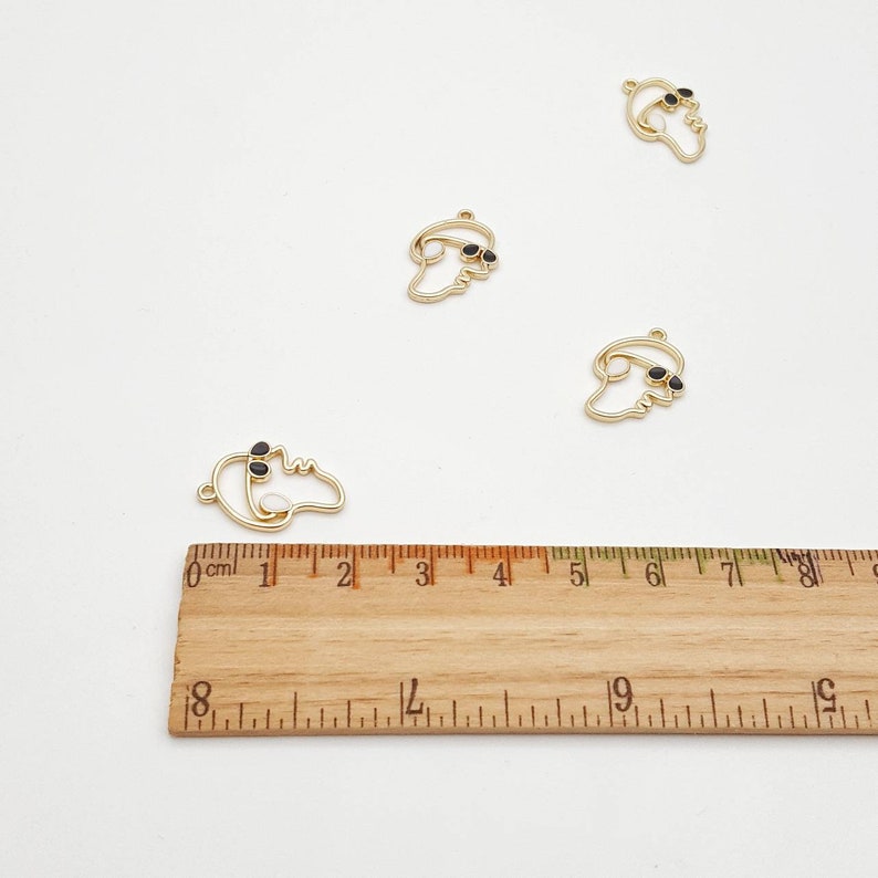 May include: Gold-colored metal charms in the shape of a face wearing sunglasses. The charms are approximately 1 inch in length. The charms are on a white surface with a wooden ruler in the foreground.