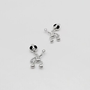 Pode incluir: Dois pingentes de esqueleto de prata com olhos pretos. Os esqueletos estão em uma pose de dança com os braços e as pernas estendidos.